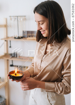 Vertical side view of happy young woman holding burning scented handmade candle in palms. Smiling female holding aromatic relaxing candle at home. Concept of wellness and relaxation. Vertical side view of happy young woman holding burning scented handmade candle in palms. Smiling female holding aromatic relaxing candle at home. Concept of wellness and relaxation. 87581062