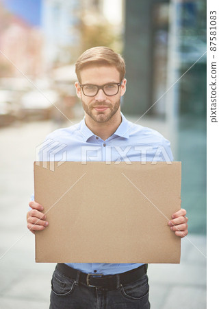 Vertical shot of a young serious man, male activist wearing blue shirt and eyeglasses holding empty sign board and looking at camera while protesting outdoors 87581083