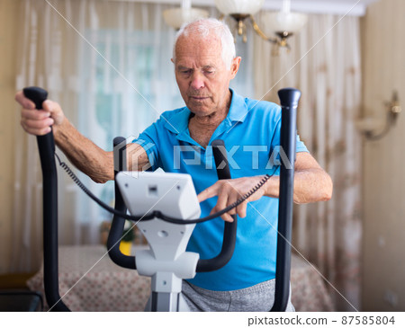 Positive senior man works out on an elliptical machine at home Positive senior man works out on an elliptical machine at home 87585804