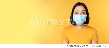 Covid-19 and medical concept. Close up portrait of asian woman in face mask, looking surprised and amazed at news, standing over yellow background Covid-19 and medical concept. Close up portrait of asian woman in face mask, looking surprised and amazed at news, standing over yellow background 87588157