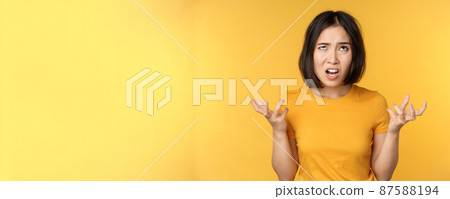 Image of angry asian woman, shouting and cursing, looking outraged, furious face expression, standing over yellow background 87588194
