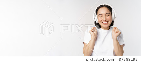 Modern asian girl dancing, listening music with headphones, smiling happy, standing in tshirt over white background 87588195