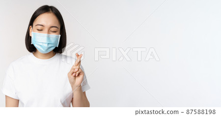 Covid-19, healthcare and medical concept. Image of asian girl in medical face mask, cross fingers, praying, making wish and smiling, standing over white background 87588198