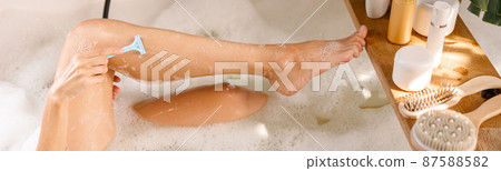 Cropped shot of young woman shaving her legs with disposable shaving razor while bathing. Body care cosmetics on wooden shelf over bathtub Cropped shot of young woman shaving her legs with disposable shaving razor while bathing. Body care cosmetics on wooden shelf over bathtub 87588582
