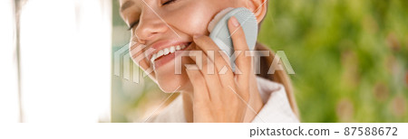 Cropped portrait of young woman wearing bathrobe smiling while using silicone face brush to cleanse skin Cropped portrait of young woman wearing bathrobe smiling while using silicone face brush to cleanse skin 87588672
