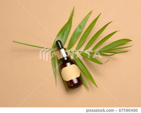 brown glass bottle with a pipette and with blank paper label stands on a brown background. Container for cosmetics, serum and oil, palm leaf 87590480