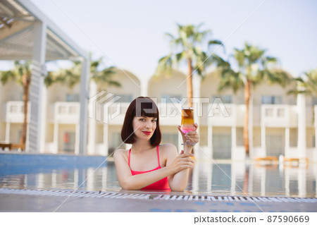 Woman in bright pink swimsuit, in resort swimming pool drinking refreshing colorful cocktail 87590669