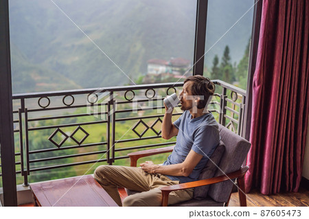 A young man holding coffee cup while sittinhg on chair on balcony , looking at mountains and green nature. Woman in Sapa in the fog, Northwest Vietnam. Vietnam travel concept. UNESCO heritage. Vietnam 87605473