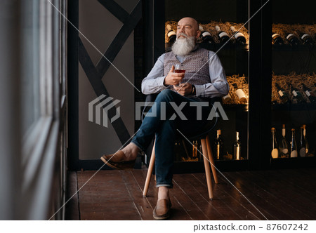 handsome man with a glass of wine sitting in a chair near a rack of collectible wine. 87607242