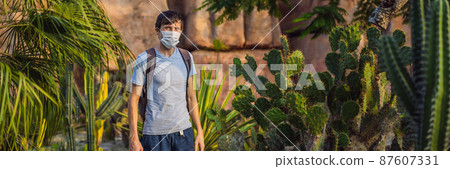 BANNER, LONG FORMAT beautiful stylish young man in desert among the cacti, wearing a medical mask during COVID-19 coronavirus, traveling in Africa on safari, exploring nature, sunny summer, traveler 87607331