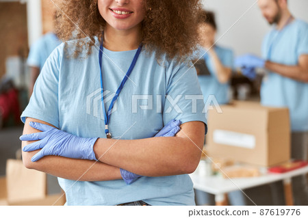 Cropped portrait of young woman, volunteer in blue uniform smiling at camera while standing with arms crossed indoors. Team sorting, packing items in the background Cropped portrait of young woman, volunteer in blue uniform smiling at camera while standing with arms crossed indoors. Team sorting, packing items in the background 87619776
