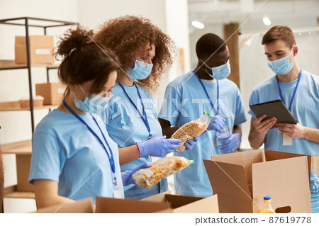 Young team of volunteers in protective masks and gloves sorting, packing food in cardboard boxes, working on donation project indoors Young team of volunteers in protective masks and gloves sorting, packing food in cardboard boxes, working on donation project indoors 87619778