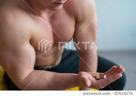 Faceless bodybuilder drinks capsules. A close-up of a muscular shirtless man holds a handful of vitamins and fitness supplements. Faceless bodybuilder drinks capsules. A close-up of a muscular shirtless man holds a handful of vitamins and fitness supplements. 87628270