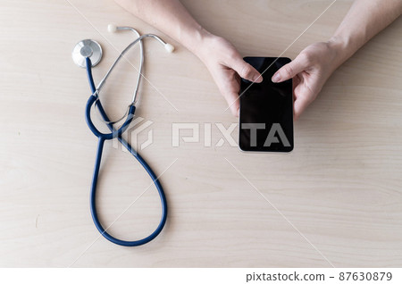 Unrecognizable female doctor sits at a desk and writes a message on a smartphone. Practitioner holds a phone in his hands and answers patients 87630879