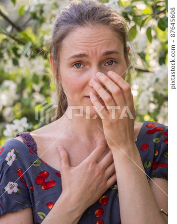 Young woman sneezes in the park against the background of a flowering tree. Allergy to pollen concept. Young woman sneezes in the park against the background of a flowering tree. Allergy to pollen concept. 87645608