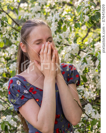 Young woman sneezes in the park against the background of a flowering tree. Allergy to pollen concept. Young woman sneezes in the park against the background of a flowering tree. Allergy to pollen concept. 87645609
