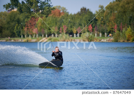 Wakeboarder surfing on lake. Young man surfer having fun wakeboarding in the cable park. Water sport, outdoor activity concept. Wakeboarder surfing on lake. Young man surfer having fun wakeboarding in the cable park. Water sport, outdoor activity concept. 87647049