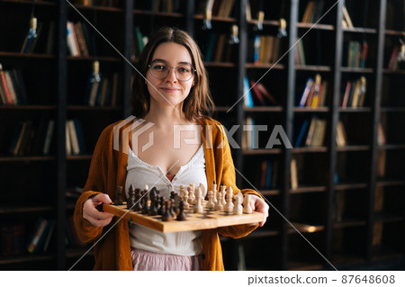 Portrait of beautiful sexy young woman in elegant eyeglasses holding in hands chess board and smiling looking at camera in dark library dark room, on blurred background of vintage bookcase. 87648608