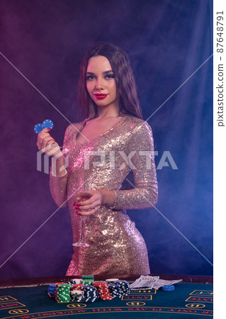 Girl playing poker at casino, holding glass of champagne, two chips. Posing at table with chips, money on it. Black, smoke background. Close-up. 87648791