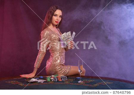 Girl playing poker at casino, holding money. Sitting on table with chips, champagne, cash on it. Black, smoke background. Close-up. 87648796