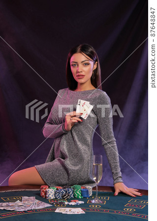 Woman in gray dress showing cards, sitting on playing table in casino. Black, smoke background with colorful backlights. Gambling, poker. Close-up. Woman in gray dress showing cards, sitting on playing table in casino. Black, smoke background with colorful backlights. Gambling, poker. Close-up. 87648847