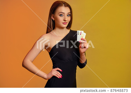 Blonde model with bright make-up, in black dress is showing two aces, posing against colorful background. Gambling, poker, casino. Close-up. Blonde model with bright make-up, in black dress is showing two aces, posing against colorful background. Gambling, poker, casino. Close-up. 87648873