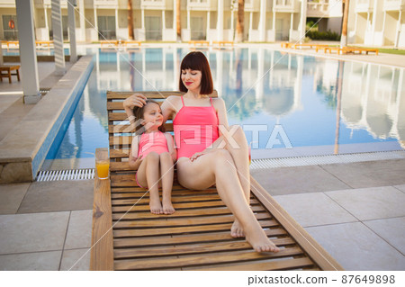 Beautiful mother holding little daughter in the same pink swimsuits while laying on deck chair 87649898