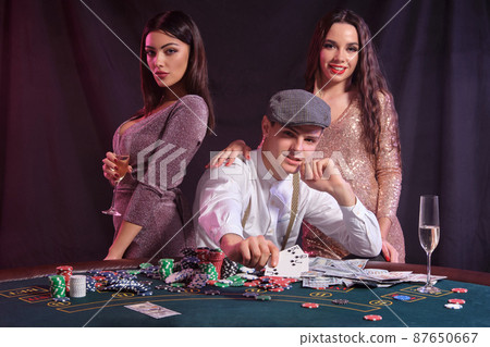Man playing poker at casino sitting at table with stacks of chips, money, cards. Celebrating win with two women. Black background. Close-up. 87650667