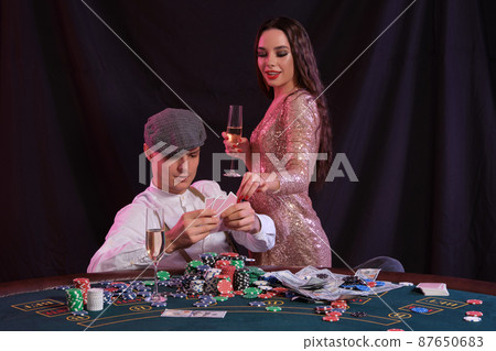 Man playing poker at casino sitting at table with stacks of chips, money, champagne, cards. Celebrating win with woman. Black background. Close-up. Man playing poker at casino sitting at table with stacks of chips, money, champagne, cards. Celebrating win with woman. Black background. Close-up. 87650683