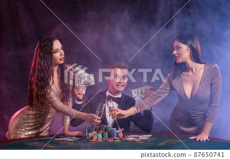 Man playing poker at casino sitting at table with stacks of chips, money, cards. Celebrating win with two women. Black, smoke background. Close-up. 87650741