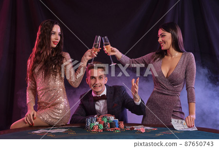 Man playing poker at casino sitting at table with stacks of chips, money, cards. Celebrating win with two women. Black, smoke background. Close-up. Man playing poker at casino sitting at table with stacks of chips, money, cards. Celebrating win with two women. Black, smoke background. Close-up. 87650743