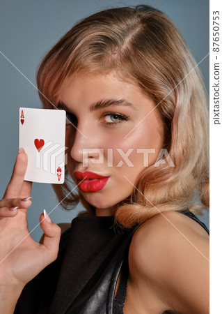 Blonde girl in black leather dress showing ace of hearts, posing against gray background. Gambling entertainment, poker, casino. Close-up. Blonde girl in black leather dress showing ace of hearts, posing against gray background. Gambling entertainment, poker, casino. Close-up. 87650753