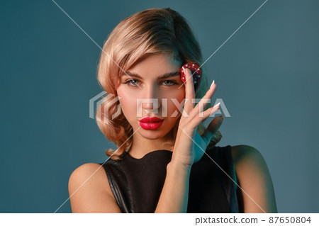 Blonde girl in black leather dress holding one red chip, posing against blue background. Gambling entertainment, poker, casino. Close-up. 87650804