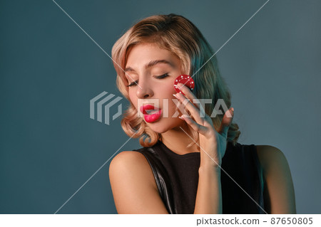 Blonde girl in black leather dress holding one red chip, posing against blue background. Gambling entertainment, poker, casino. Close-up. Blonde girl in black leather dress holding one red chip, posing against blue background. Gambling entertainment, poker, casino. Close-up. 87650805