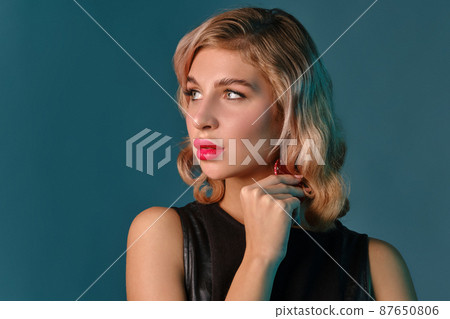 Blonde girl in black leather dress showing two red chips, posing against blue background. Gambling entertainment, poker, casino. Close-up. 87650806