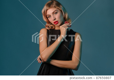 Blonde girl in black leather dress showing two red chips, posing against blue background. Gambling entertainment, poker, casino. Close-up. 87650807