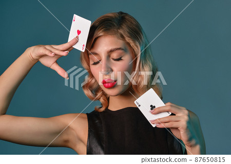 Blonde girl in black leather dress showing two playing cards, posing against blue background. Gambling entertainment, poker, casino. Close-up. Blonde girl in black leather dress showing two playing cards, posing against blue background. Gambling entertainment, poker, casino. Close-up. 87650815