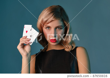 Blonde girl in black stylish dress showing two playing cards, posing against blue background. Gambling entertainment, poker, casino. Close-up. 87650816
