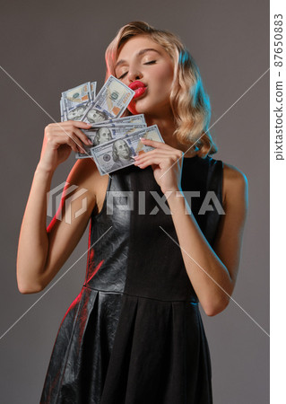 Blonde girl in black stylish dress holding and kissing some money, posing against gray background. Gambling entertainment, poker, casino. Close-up. Blonde girl in black stylish dress holding and kissing some money, posing against gray background. Gambling entertainment, poker, casino. Close-up. 87650883