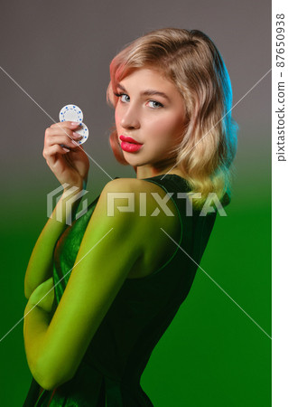 Blonde girl in black stylish dress holding two chips, posing against gray background, green flare. Gambling entertainment, poker, casino. Close-up. Blonde girl in black stylish dress holding two chips, posing against gray background, green flare. Gambling entertainment, poker, casino. Close-up. 87650938