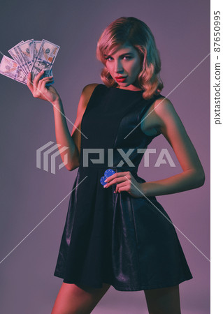 Blonde girl in black stylish dress holding some money and chips, posing against colorful background. Gambling entertainment, poker, casino. Close-up. 87650995