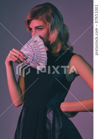Blonde girl in black stylish dress holding some money, posing against colorful background. Gambling entertainment, poker, casino. Close-up. 87651001