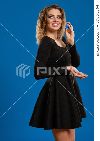 Blonde maiden in black short dress is smiling, showing colorful chips, posing sideways on blue background. Gambling, poker, casino. Close-up 87651364