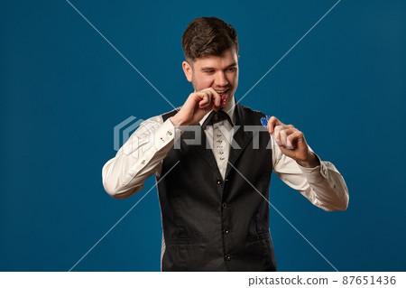 Noob in poker, in black vest and white shirt. Holding some colored chips. Posing against blue background. Gambling, casino. Close-up. Noob in poker, in black vest and white shirt. Holding some colored chips. Posing against blue background. Gambling, casino. Close-up. 87651436