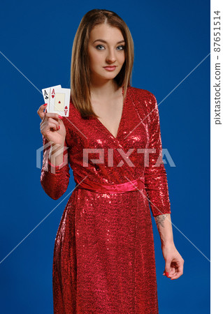Blonde woman with make-up, tattooed hand, in red sequin dress is showing two aces, posing on blue background. Gambling, poker, casino. Close-up. Blonde woman with make-up, tattooed hand, in red sequin dress is showing two aces, posing on blue background. Gambling, poker, casino. Close-up. 87651514