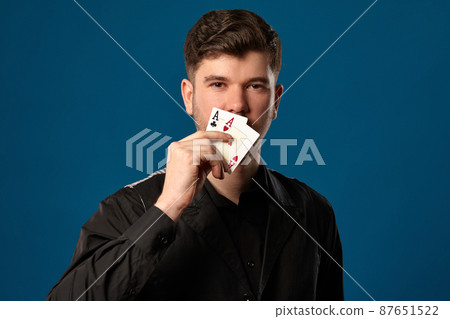 Newbie in poker, in black vest and shirt. Holding two playing cards while posing against blue studio background. Gambling, casino. Close-up. Newbie in poker, in black vest and shirt. Holding two playing cards while posing against blue studio background. Gambling, casino. Close-up. 87651522