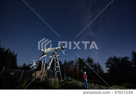 Male worker pointing hand at pump jack under beautiful night sky with stars. Petroleum engineer controlling work of oil pump rocker-machine at night in oil field. 87651589