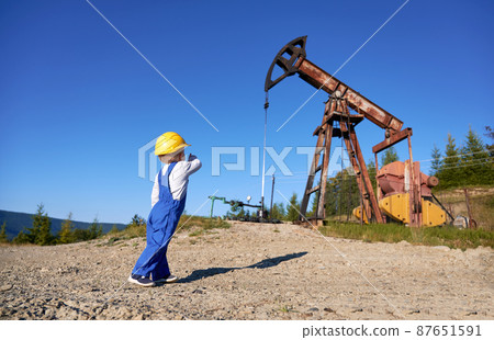 Back view of little boy in construction helmet who standing nearby rocking pump for oil extraction. Male child pointing to petroleum rig while standing on stone road at summer day. Back view of little boy in construction helmet who standing nearby rocking pump for oil extraction. Male child pointing to petroleum rig while standing on stone road at summer day. 87651591
