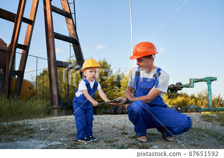 Engineer father introducing his son to work tool. Adult man and preschool child in workwears and helmets together looking to adjustable wrench on background of pump for oil production. Engineer father introducing his son to work tool. Adult man and preschool child in workwears and helmets together looking to adjustable wrench on background of pump for oil production. 87651592