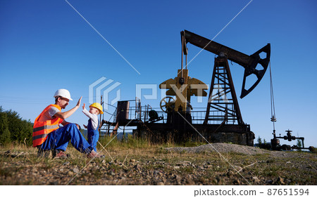 Adult man and small boy in work uniforms and construction helmets clapping their hands each other. On the background oil rig with rocking pump under blue sky. Adult man and small boy in work uniforms and construction helmets clapping their hands each other. On the background oil rig with rocking pump under blue sky. 87651594